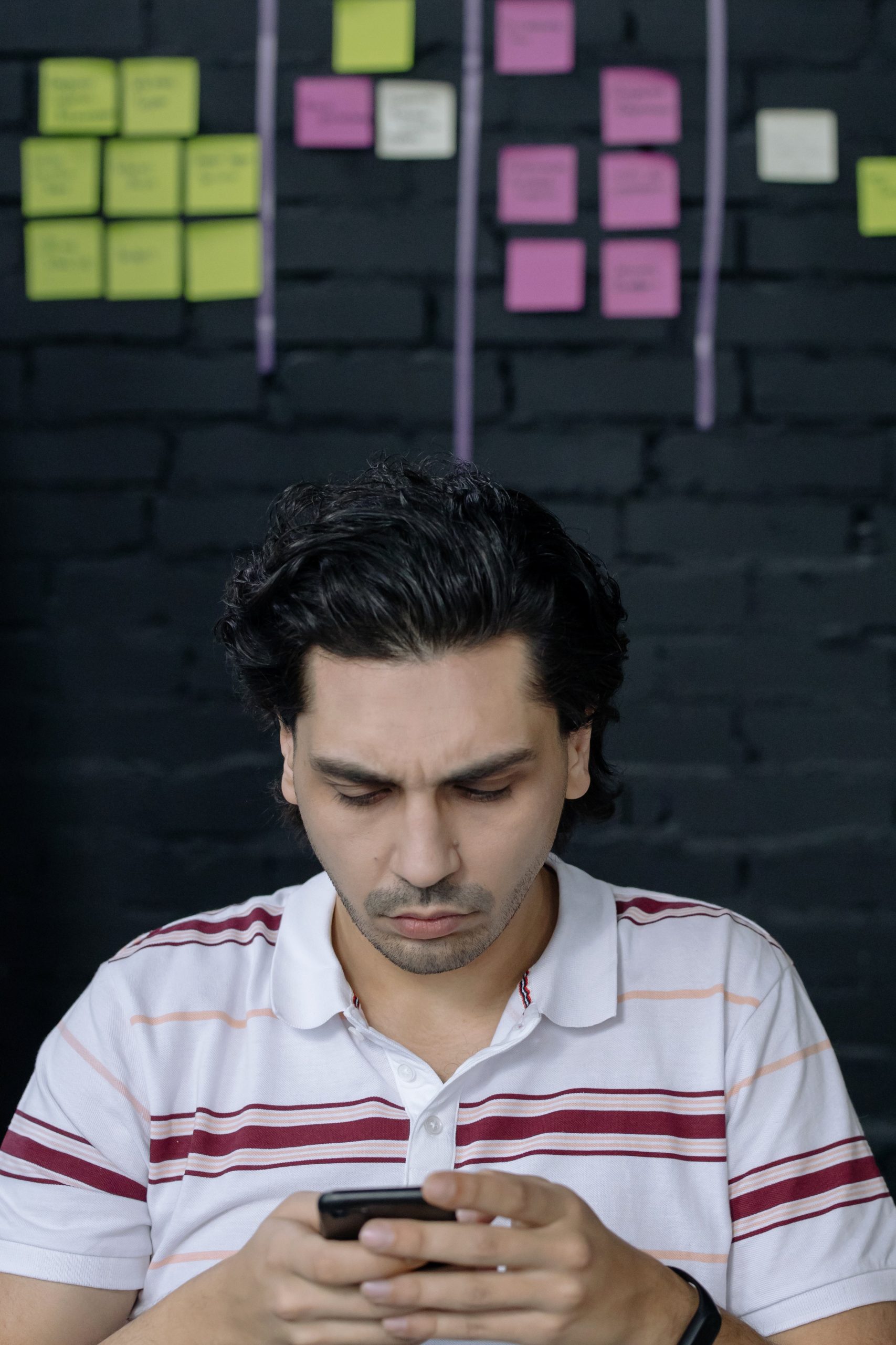 guy wearing white polo shirt with stripes, looking intently at phone. With different colored sticky notes on the wall behind him